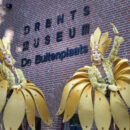 Twee steltenlopers die verkleed zijn als bloemen zwaaien naar de camera met het logo van het museum op de achtergrond