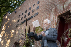 Man houdt een toespraak met het logo van Drents Museum De Buitenplaats op de achtergrond