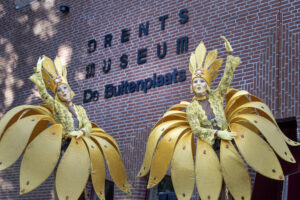 Twee steltenlopers die verkleed zijn als bloemen zwaaien naar de camera met het logo van het museum op de achtergrond