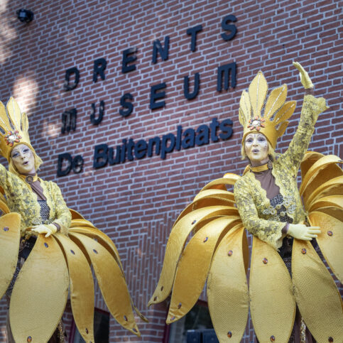Twee steltenlopers die verkleed zijn als bloemen zwaaien naar de camera met het logo van het museum op de achtergrond